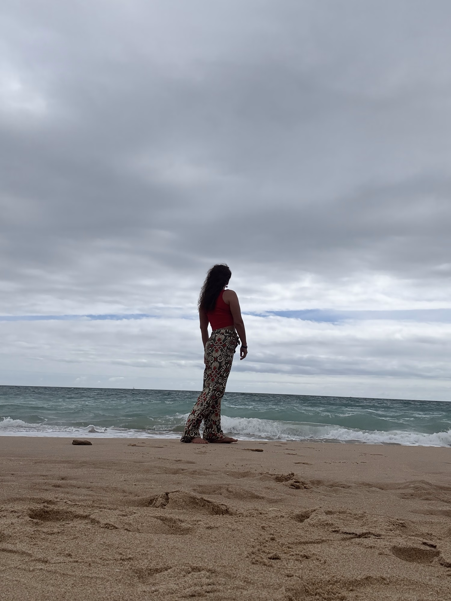 A person in a red top and patterned trousers stands on a sandy beach, looking out at the sea beneath a cloudy sky.
