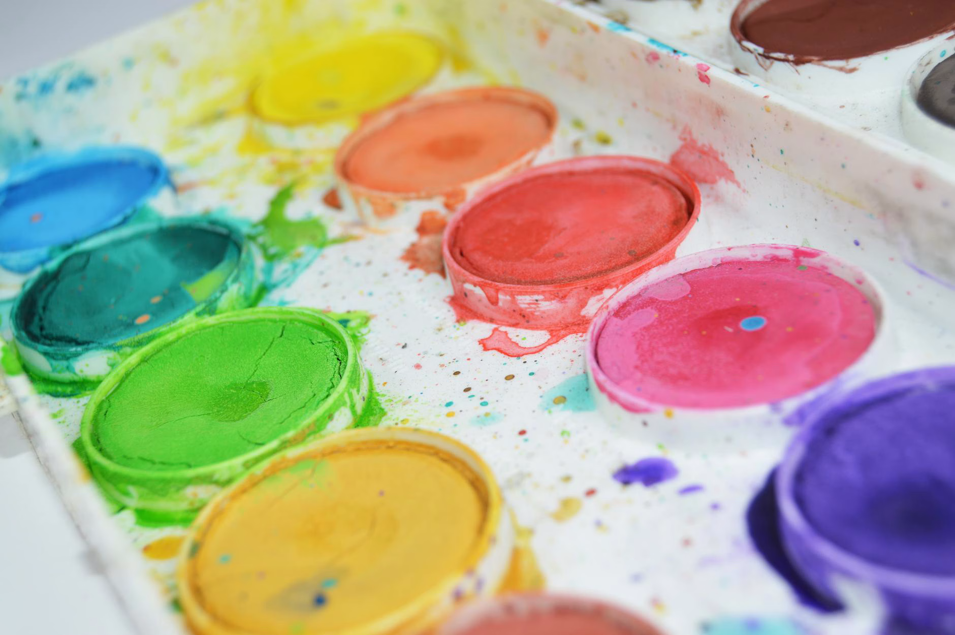 A close-up shows a watercolour palette with bright, used paints and colourful splatters on the white tray.