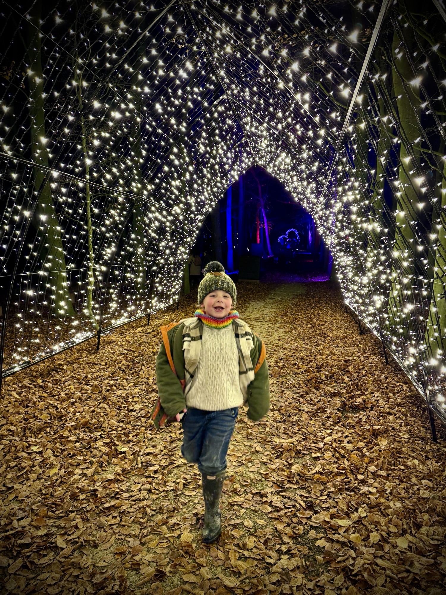 A child wearing wellies and a winter hat walks at night through a tunnel of white lights on a leafy path.