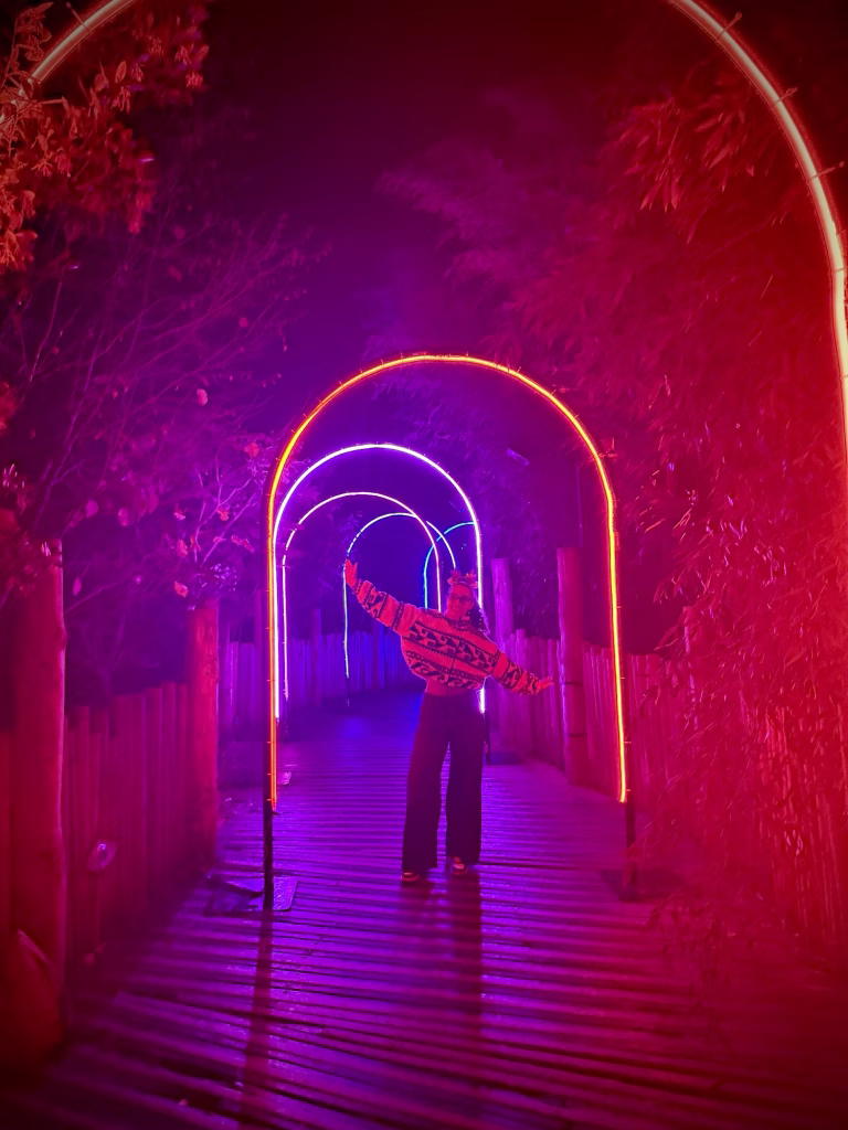 A person stands on a wooden walkway at night beneath glowing neon arches, arms outstretched in red and purple light.