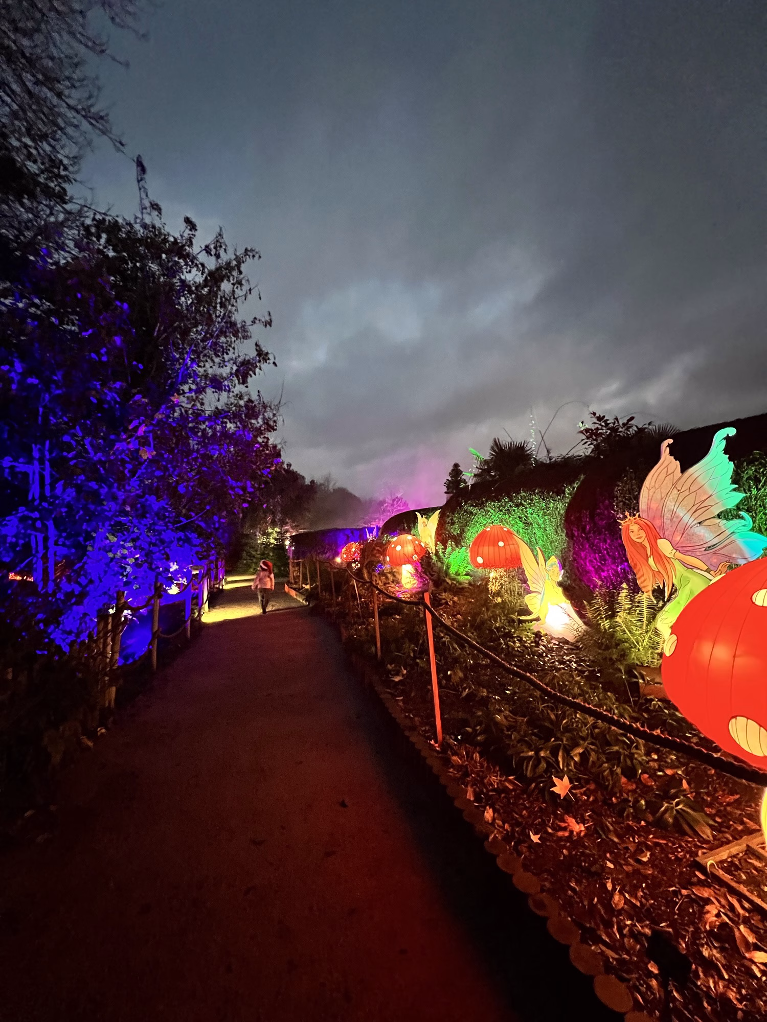At dusk, a person walks along a path decorated with glowing lanterns and plants beneath a cloudy sky.