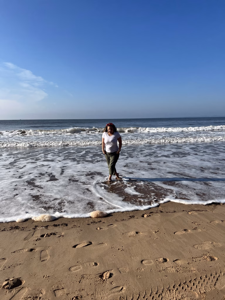 Someone stands by the shore as gentle waves near their feet, leaving footprints in the sand beneath a clear blue sky.