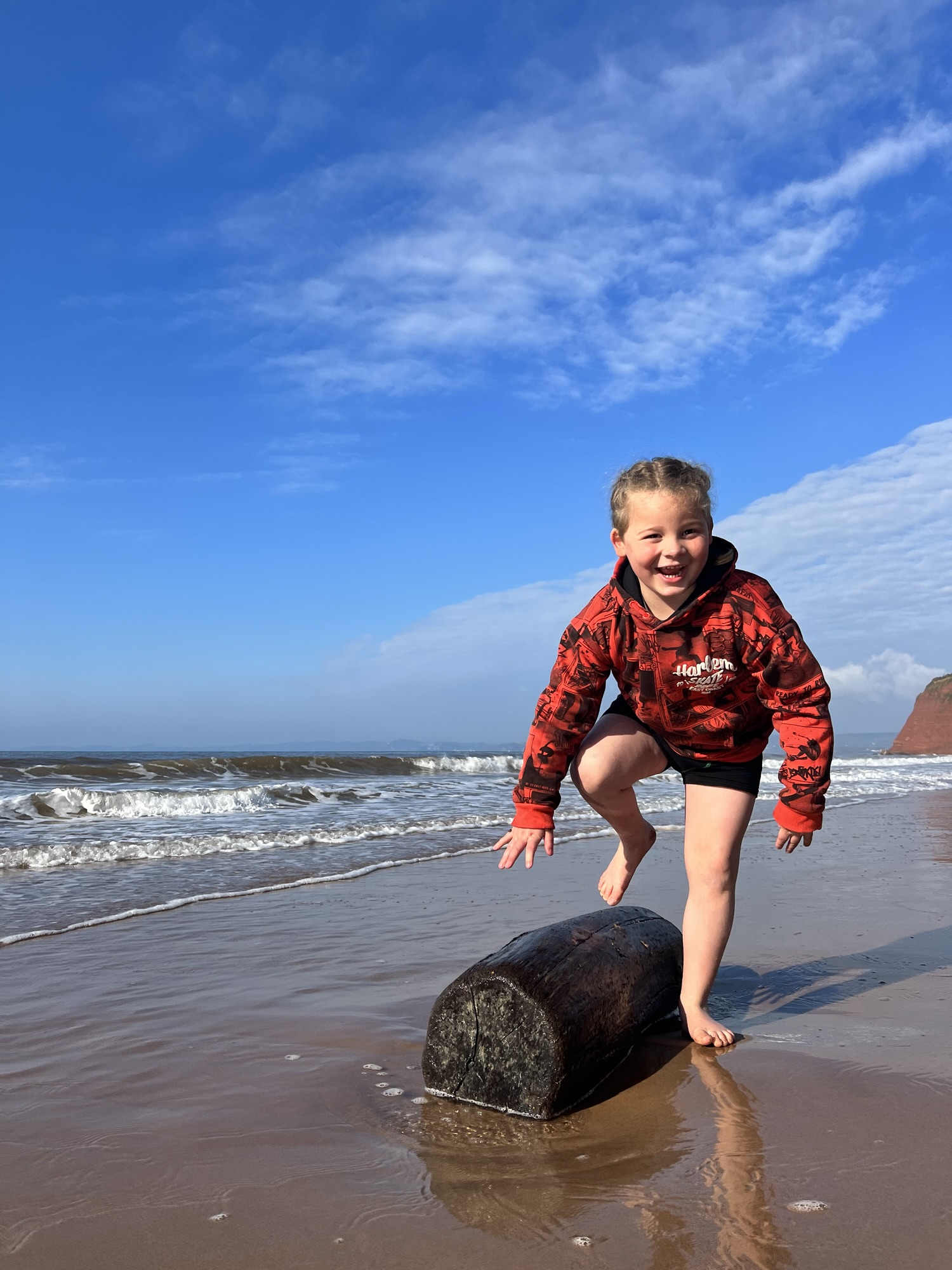 A smiling child in a red hoodie stands on a driftwood log at the beach, with waves and blue sky behind them.