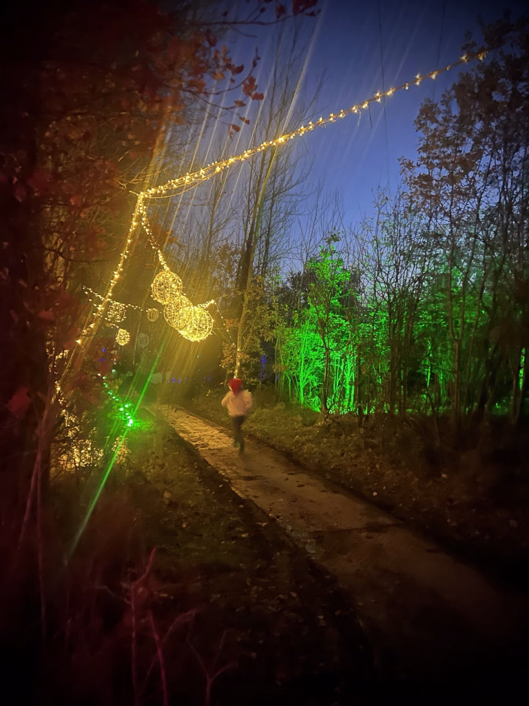 At dusk, a child runs down a narrow path lined with trees glowing in green and yellow, with festive lights hanging above.
