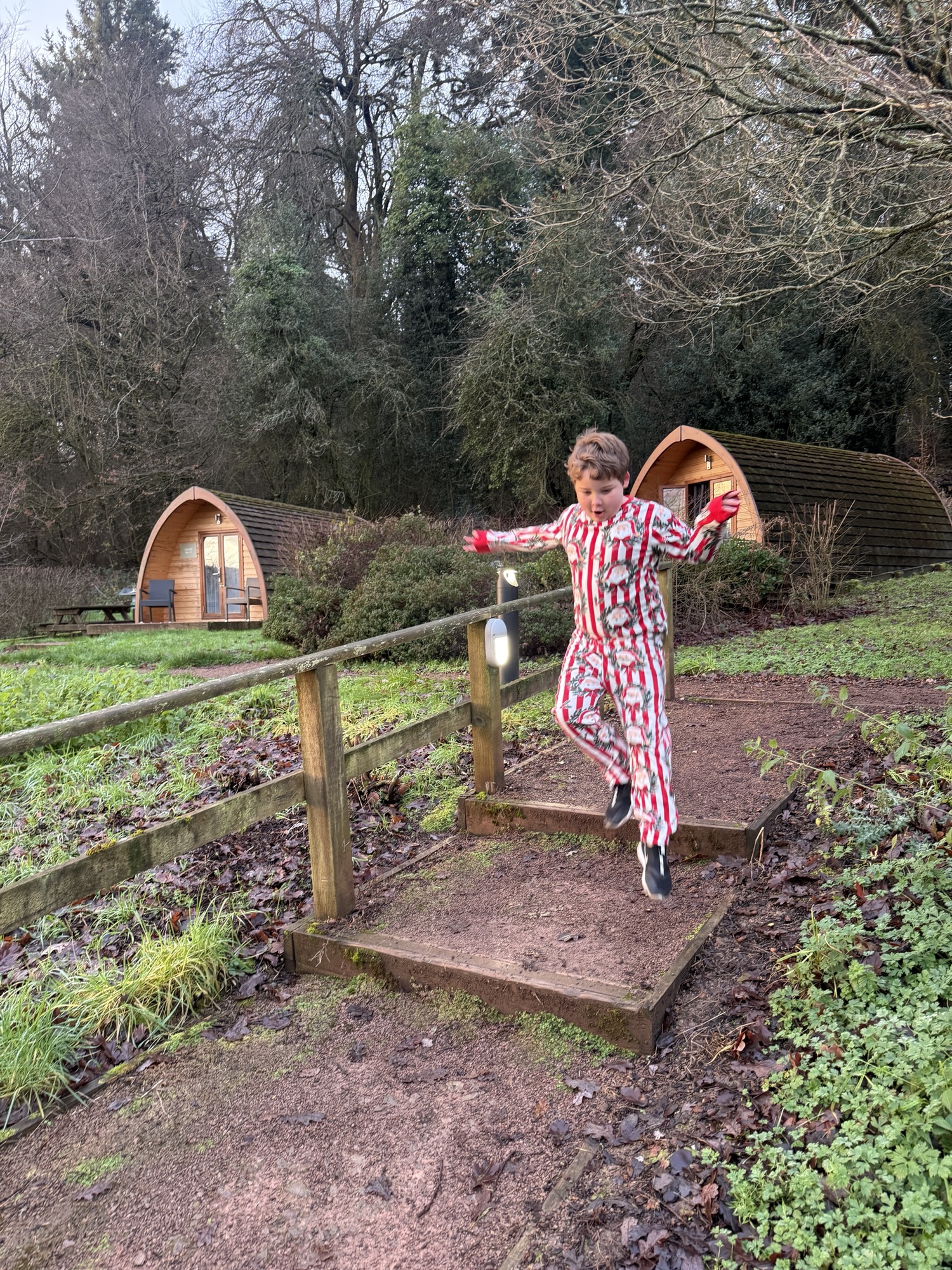 Someone in a patterned jumpsuit leaps off a small wooden bridge near cabins surrounded by grass and trees.