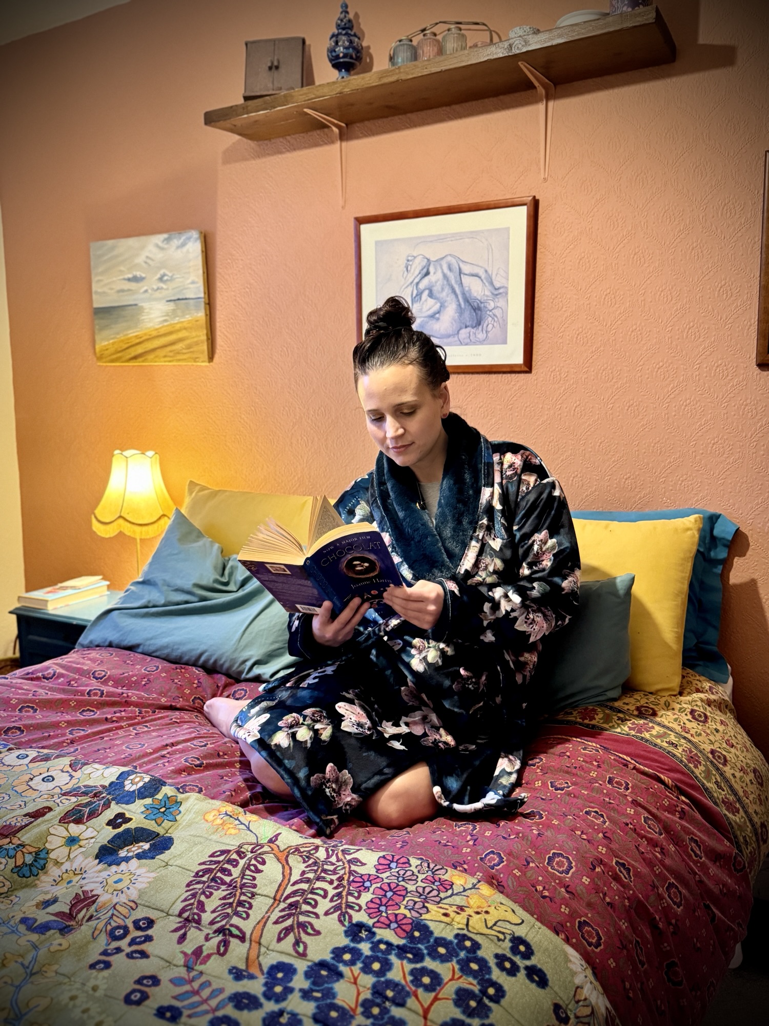 Someone in a floral dressing gown reads on a colourful bed in a warmly lit, cosy bedroom decorated with artwork.