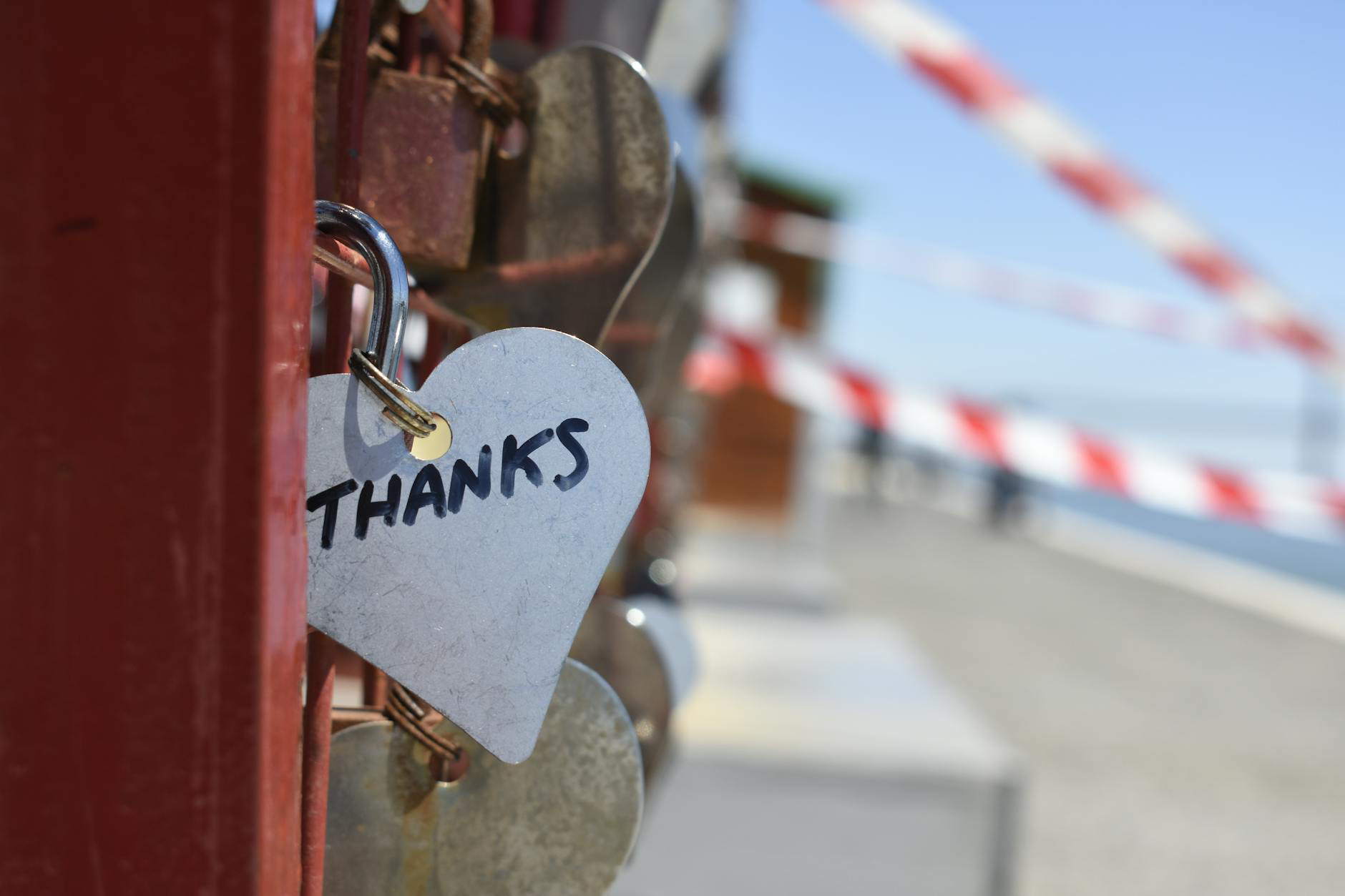 A heart-shaped padlock labelled "THANKS" hangs on a fence with other locks, against a blurred background and red-and-white tape.