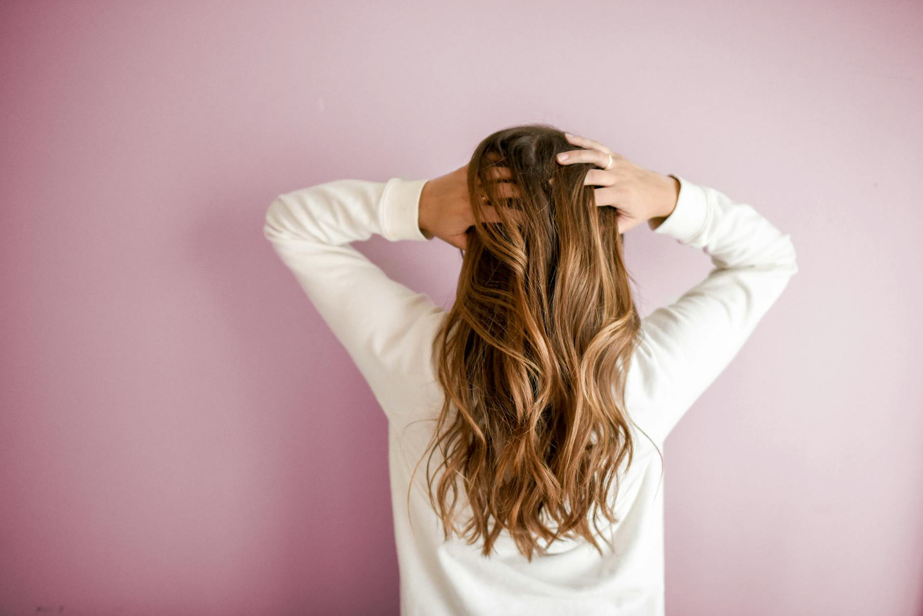 A person with long, wavy brown hair stands by a pink wall in a white top, holding their head with both hands.