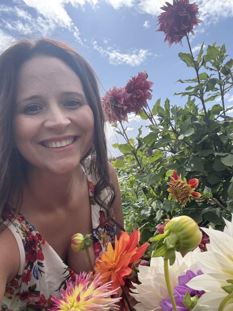 A smiling woman in a floral dress stands surrounded by colourful flowers beneath a sky with scattered clouds.