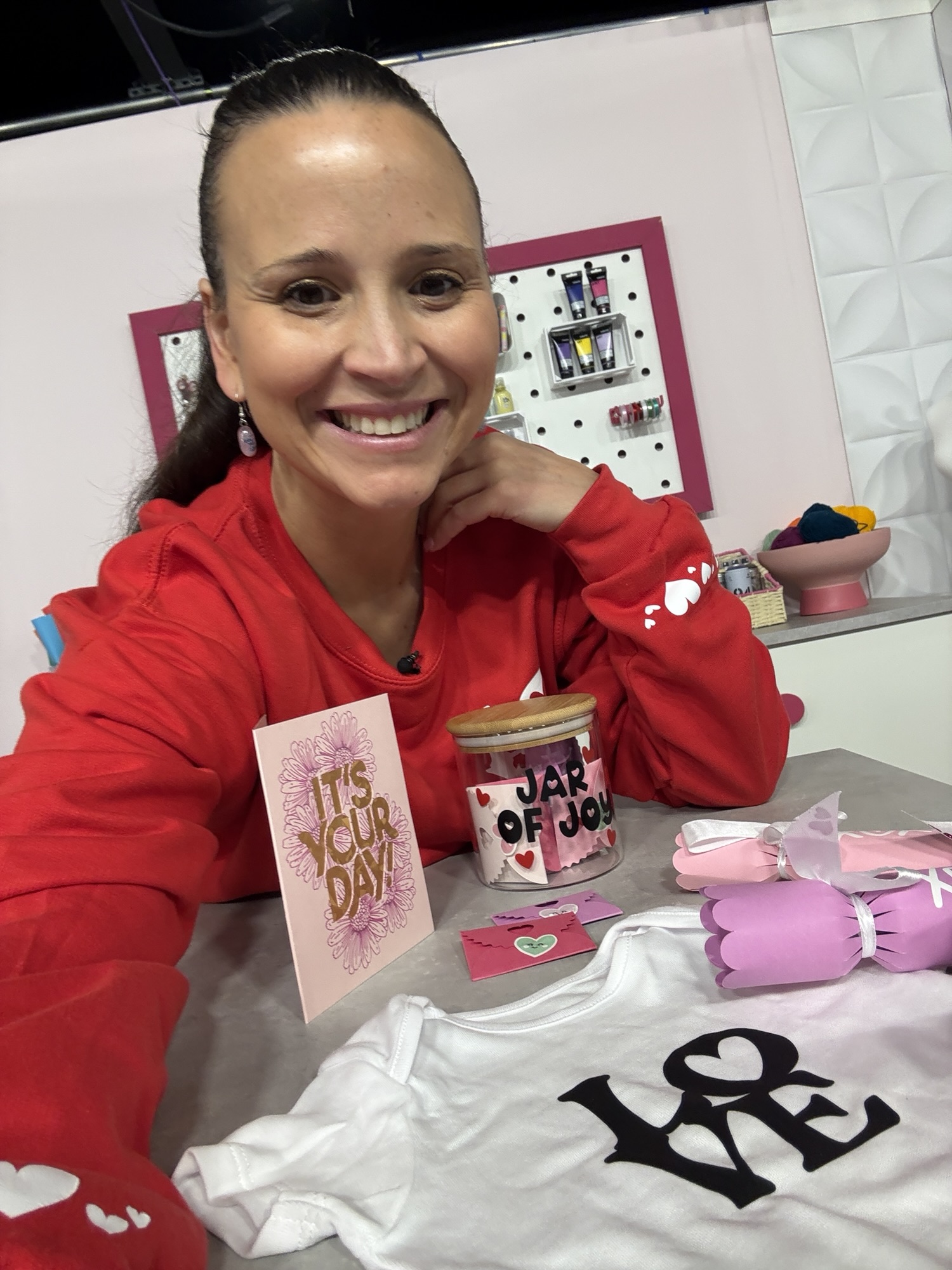A smiling woman in a red jumper sits at a table with a card, Jar of Joy, pink gloves, and a baby vest reading "LOVE.