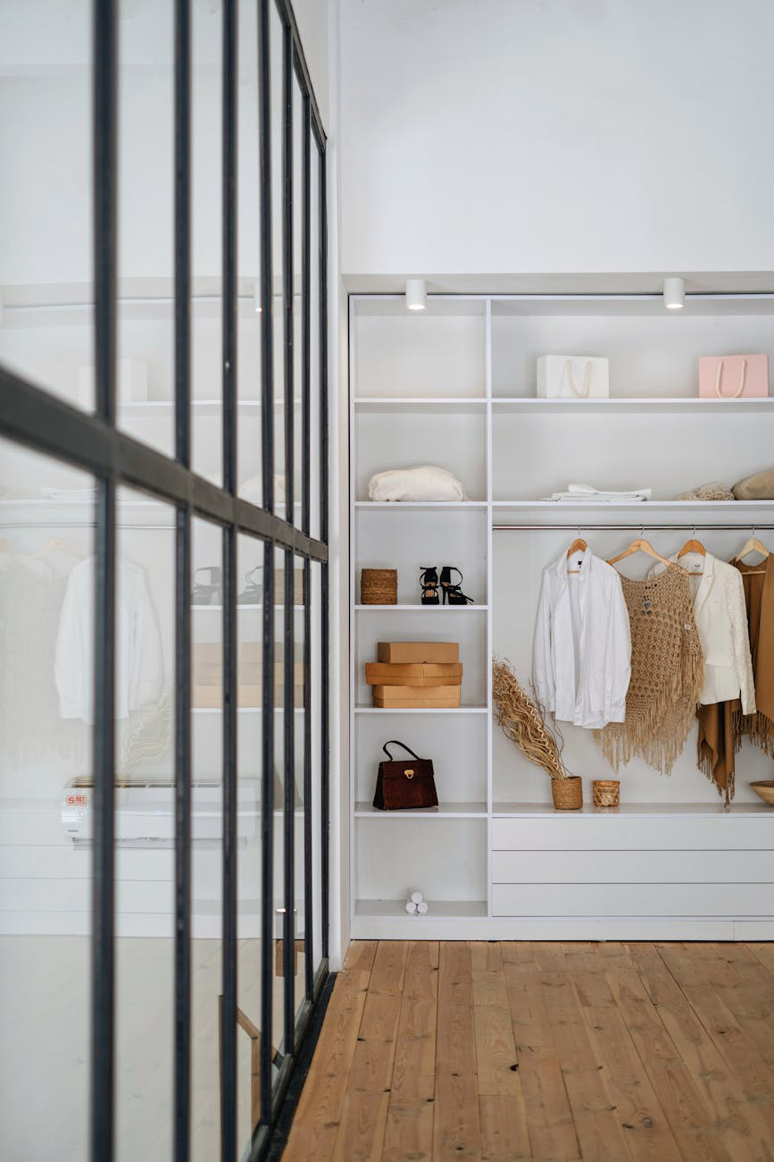 A tidy walk-in wardrobe with open white shelves holds clothes and bags, beside a glass wall and wooden floor.