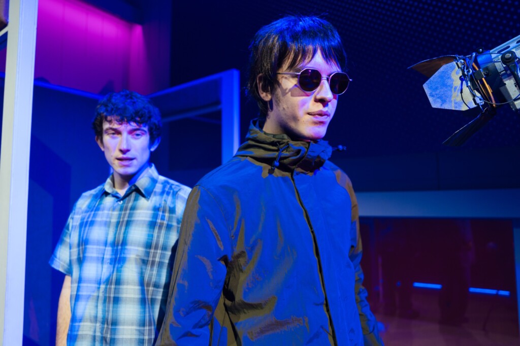 Two men stand under stage lights; one is in sunglasses and a dark jacket, the other in a checked shirt.