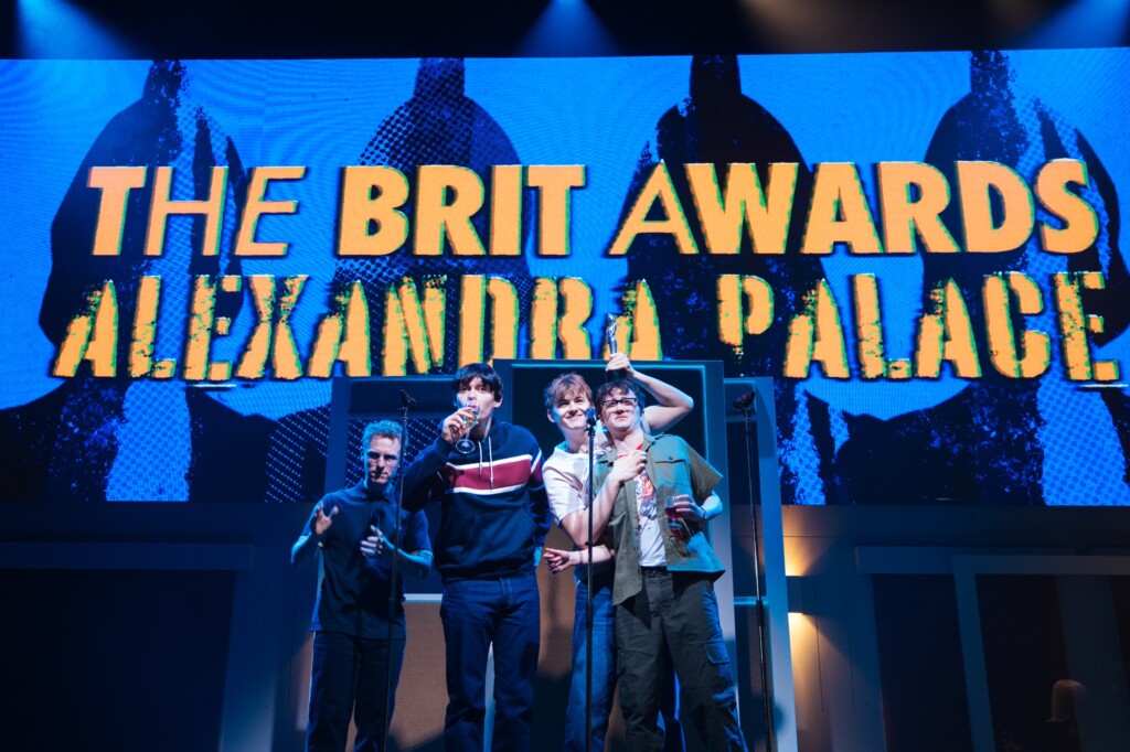Four people stand on stage with microphones and drinks, in front of a big screen showing "The Brit Awards Alexandra Palace.