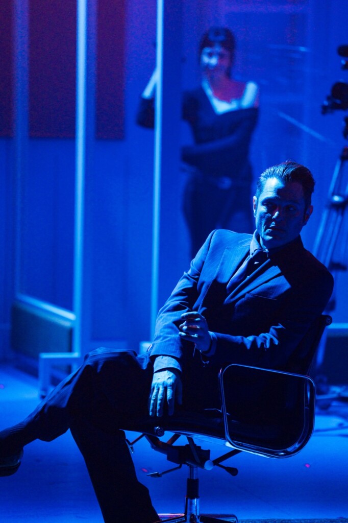 A man in a suit sits in an office chair under blue lights, with a woman visible behind glass panels in the background.