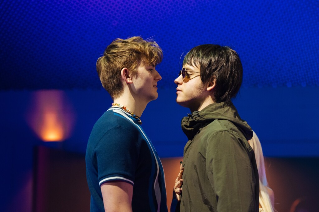 Two young men stand closely facing each other in a room lit with soft blue and purple light.
