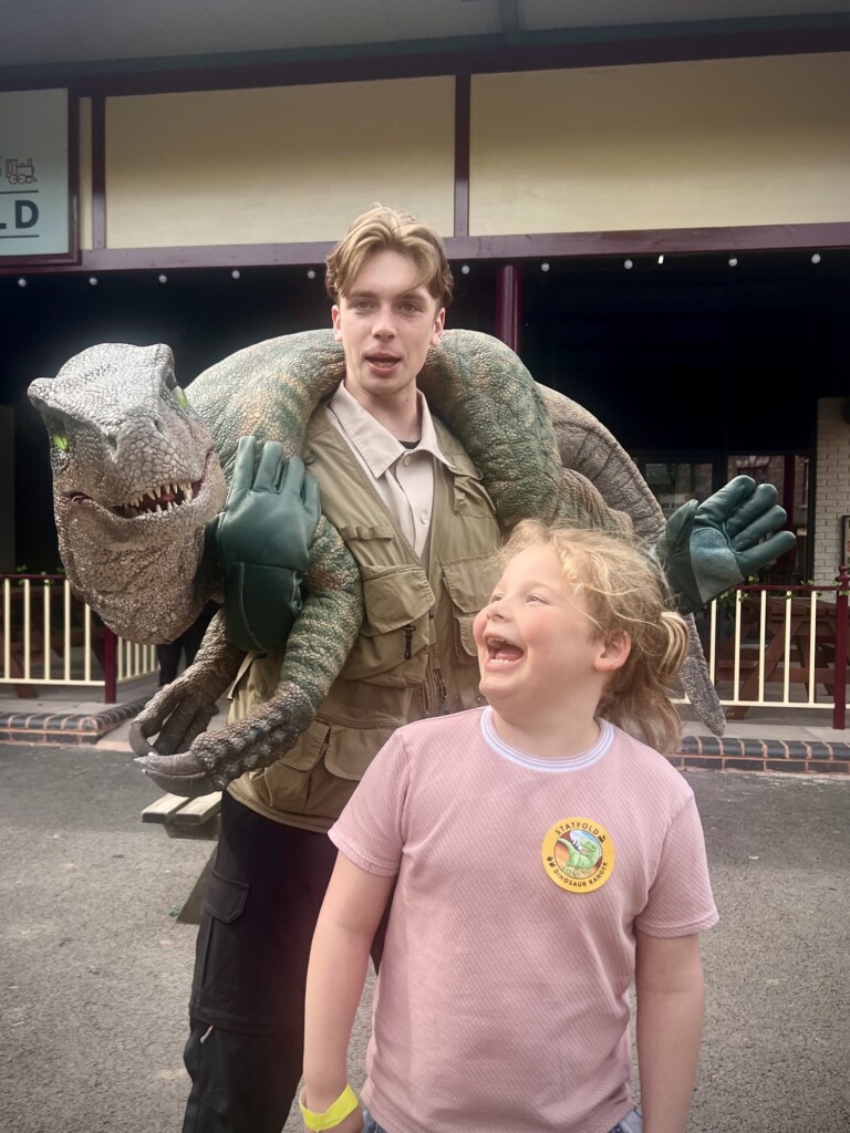A man holding a lifelike dinosaur puppet stands behind a smiling child in a pink shirt with a sticker, outdoors.