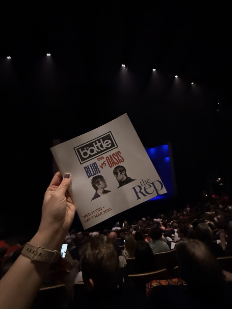 Someone holds up a "The Battle: Blur vs Oasis" leaflet over an audience in a dimly lit theatre auditorium.