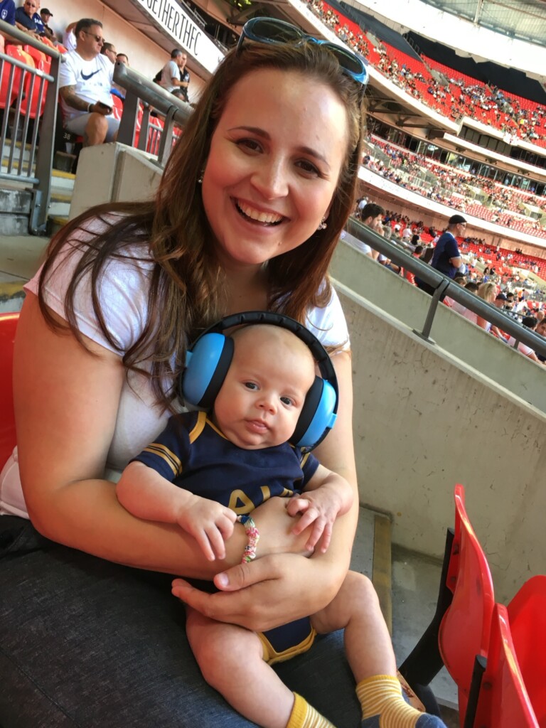 A woman holds a baby in noise-cancelling headphones and smiles at a stadium event.