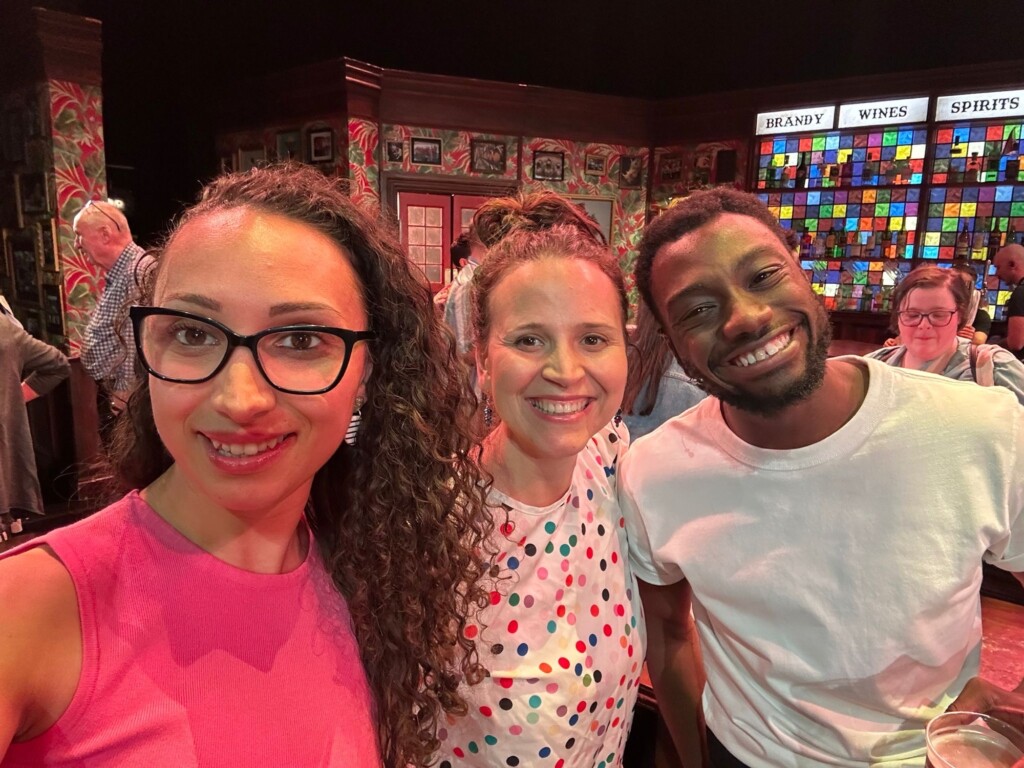 Three friends take a selfie together in a bright room with stained glass and shelves filled with bottles behind them.