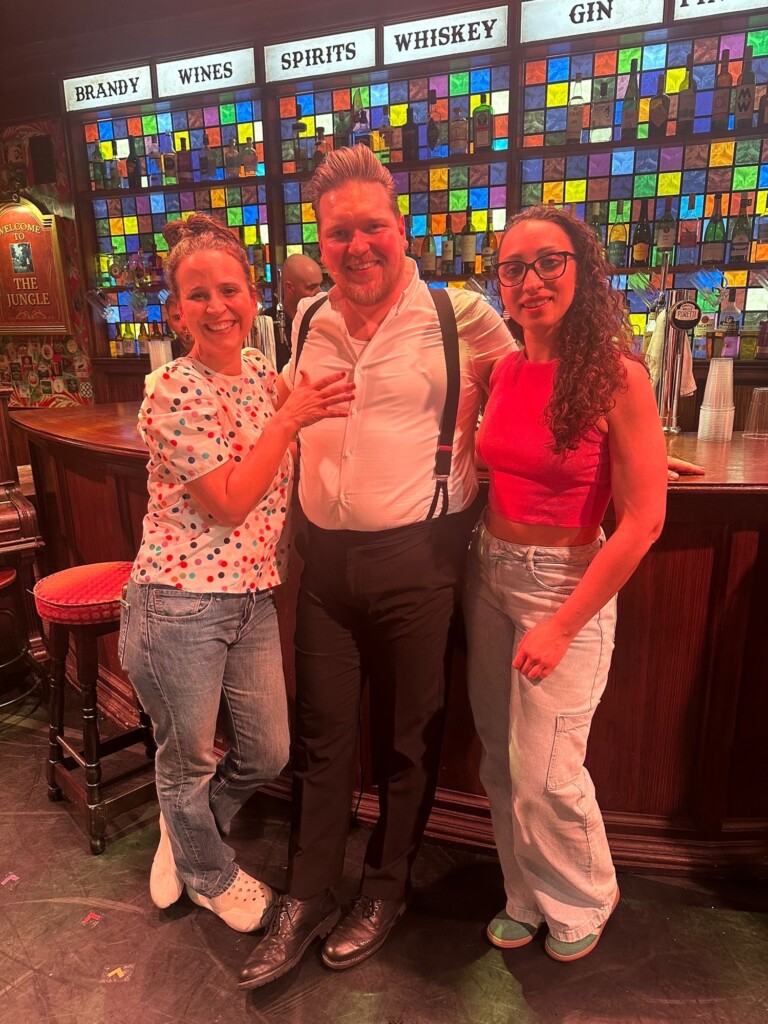 Three smiling people, two women and a man in braces, pose together in front of a colourful bar.
