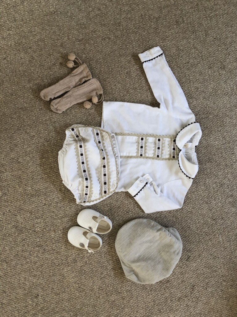 A baby outfit with a white shirt, matching shorts, beige pom-pom socks, white shoes, and a grey cap is laid out on brown carpet.