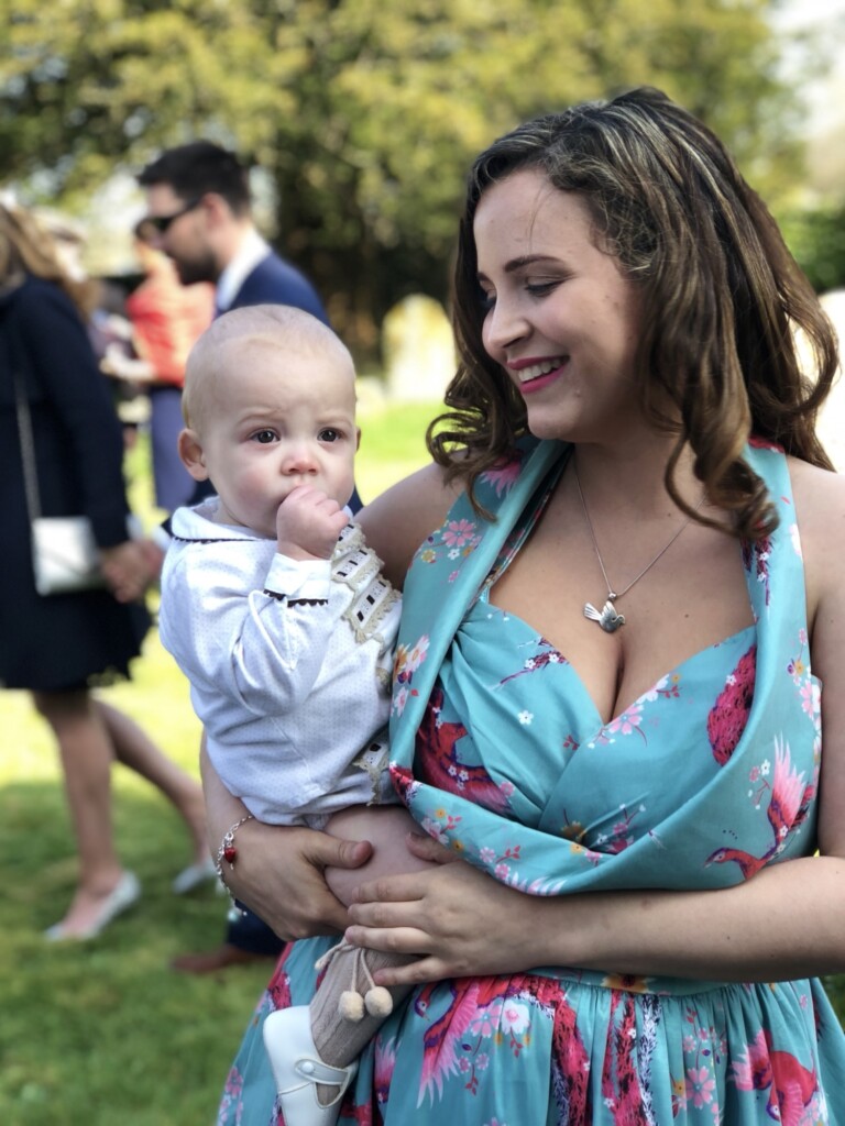 A woman in a floral dress holds a smiling baby outdoors, with several people blurred in the sunny background.