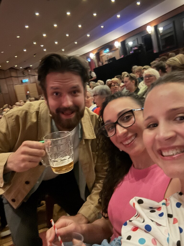 In a crowded theatre, a man with a beer tankard leans towards the camera as two women beside him smile.