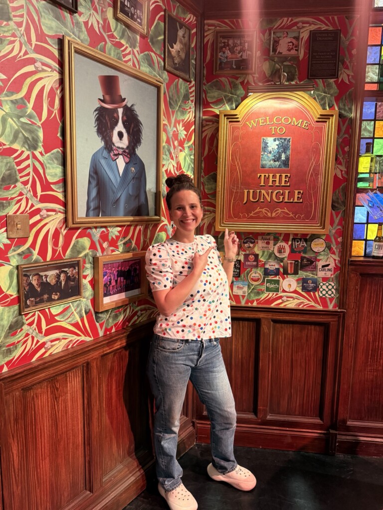 Someone smiles and points at a "Welcome to the Jungle" sign in a colourful room with photos and a dog portrait in a suit.