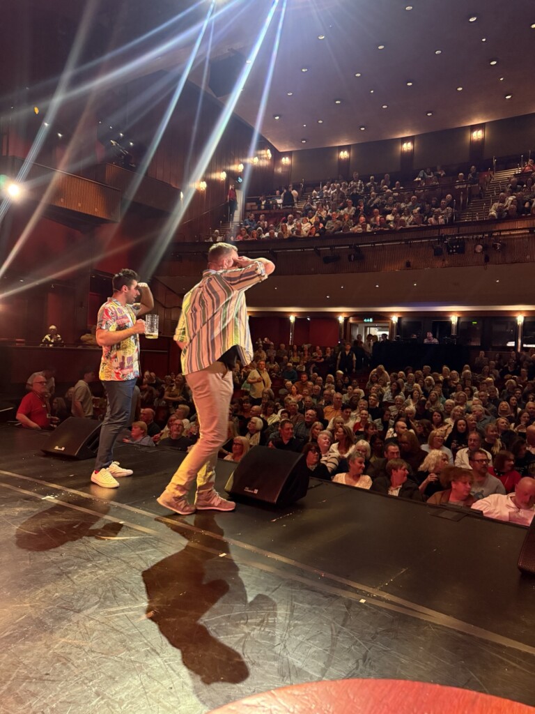 Two performers face a full theatre audience; one drinks from a bottle while the other shades his eyes from the stage lights.