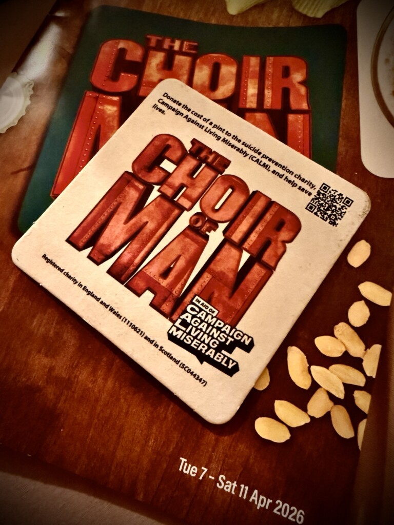 A coaster with "CHOIR OF MAN" sits on a show programme dated 7–11 April 2026, with some snacks nearby.