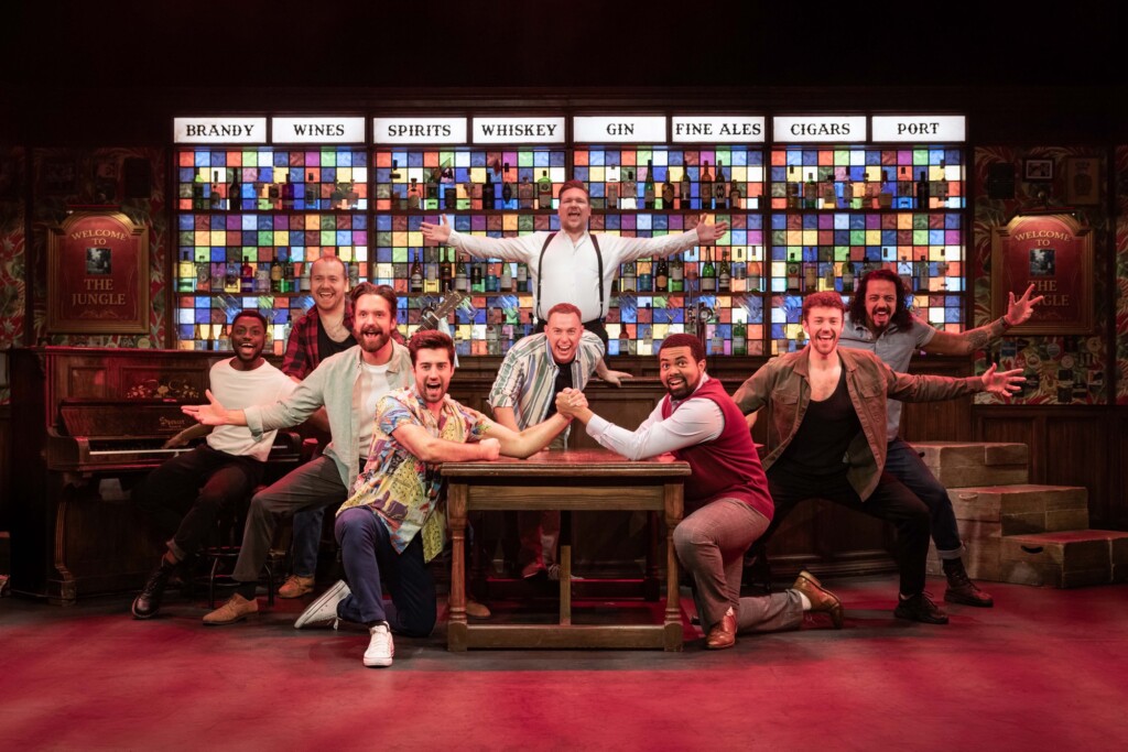 Nine men pose around a table on stage, with a bright drinks menu visible behind them.