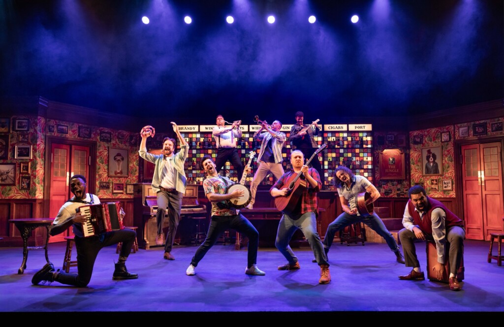 Nine musicians play instruments on a vibrant, well-lit stage that looks like a bustling bar or club.