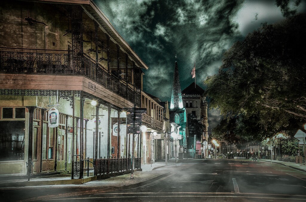 A quiet night-time street with old buildings and glowing windows, and a church spire rising against the cloudy sky.