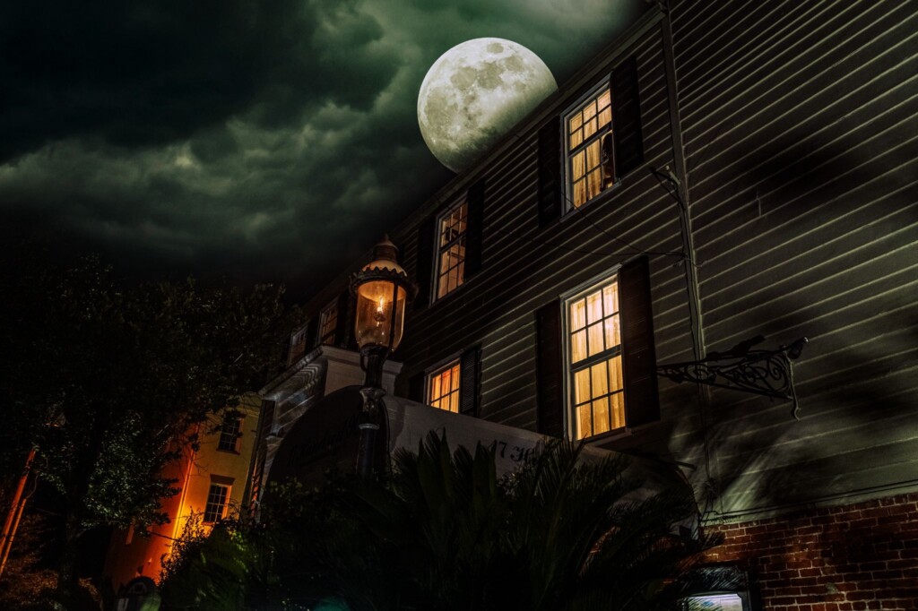 A bright full moon illuminates a dark wooden building with glowing windows and an old streetlamp on a cloudy night.
