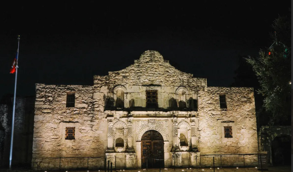 The Alamo’s stone front is lit up at night, with a flagpole standing to the left side of the building.