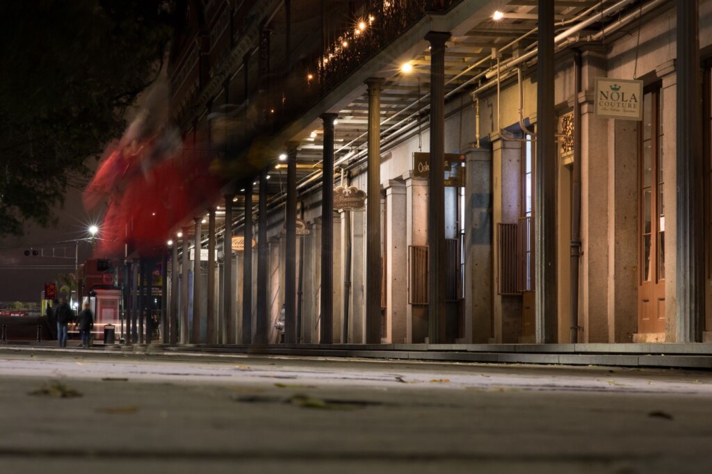 A New Orleans building with columns and open shutters at night, featuring a NOLA Couture sign as people walk past on the pavement.