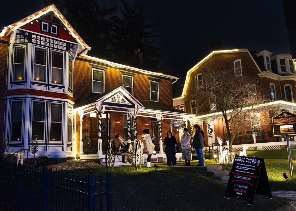 People stand outside a brick house lit by white Christmas lights at night, with a ghost tour sign in the garden.