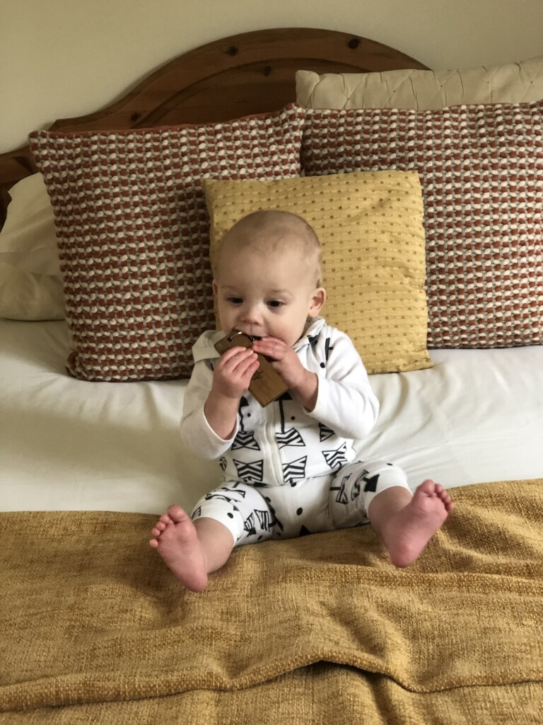A baby in patterned clothes sits on a bed, playing and chewing on a brown object among yellow and patterned cushions.