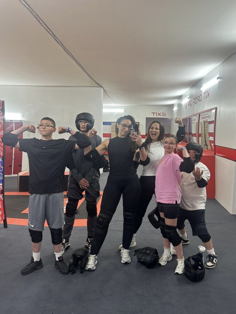 Five people in helmets and sportswear pose and flex in a gym, standing by a mirror against red and white walls.
