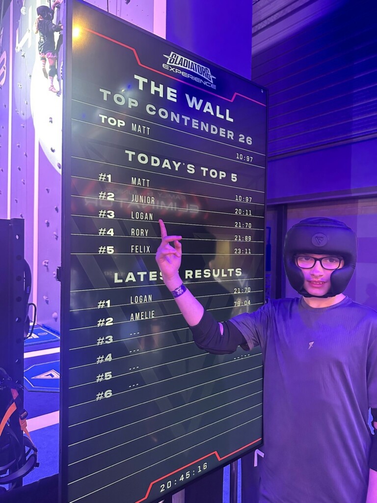 A person in a helmet and glasses points at a leaderboard showing the top five contenders for "The Wall" climbing challenge.