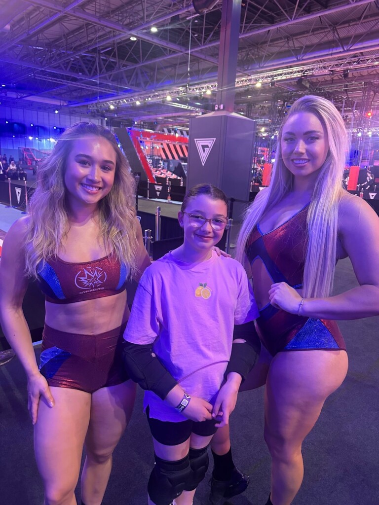 Two women in sports outfits stand on each side of a child with glasses and a purple shirt in a large indoor venue.
