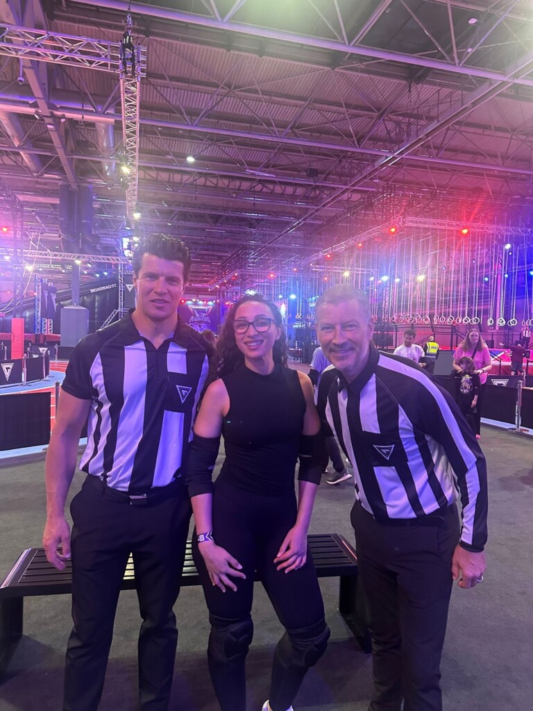 Two referees and a woman in sportswear stand together indoors, with obstacle course equipment behind them.