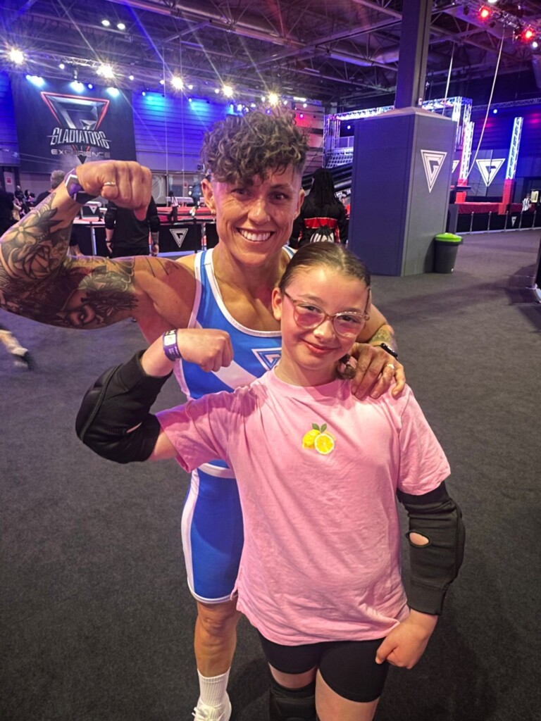 An athlete and a smiling child both flex their arms together at an indoor event.