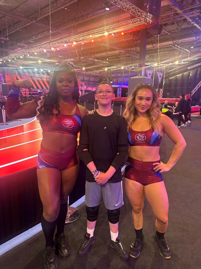A boy stands between two women in sportswear at an indoor event, with a red-lit arena visible behind them.