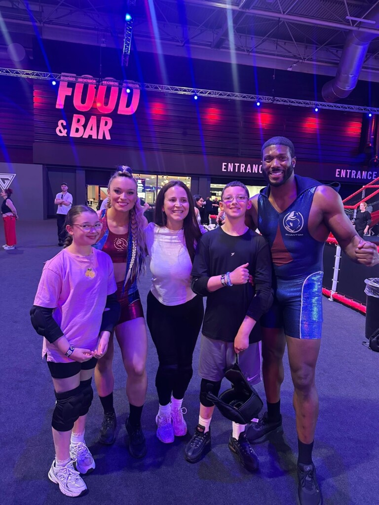 Two adults in sportswear and three children pose together at an indoor event, with signs for food, bar, and entrance behind them.