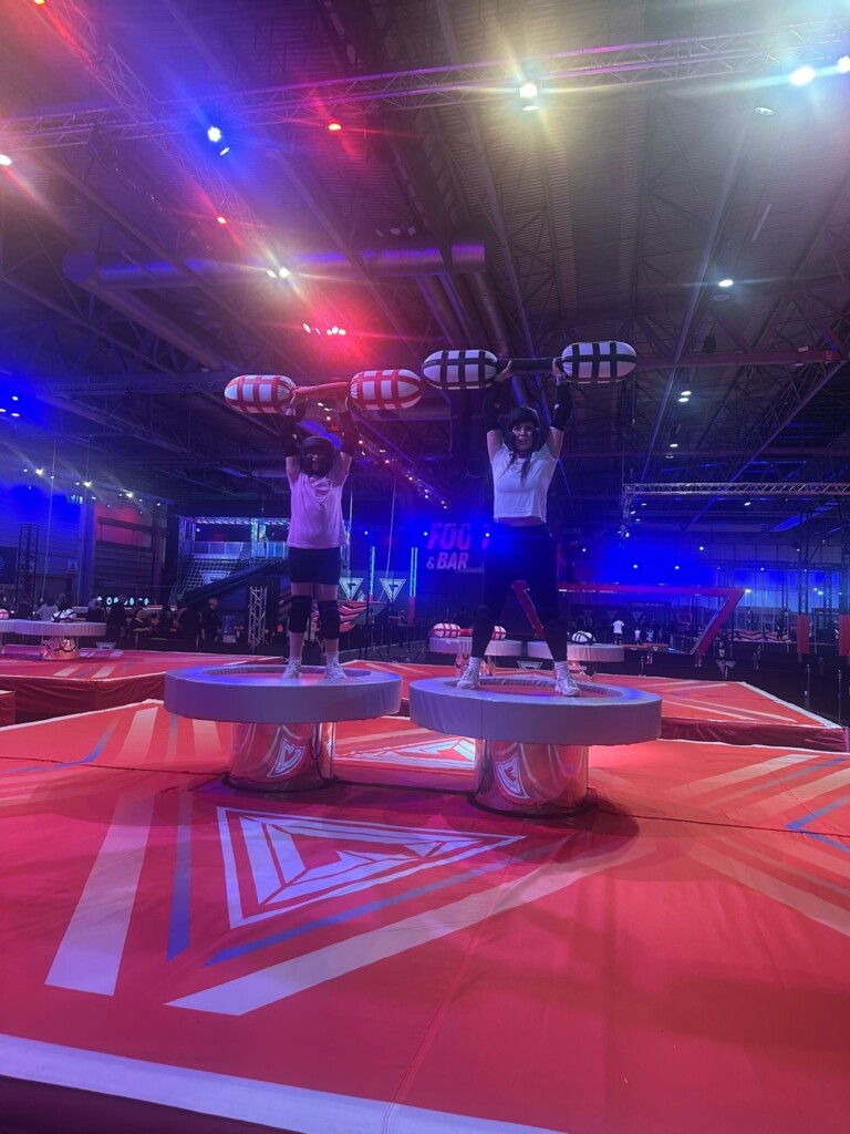 Two people balance on round platforms indoors, raising padded jousting sticks under red and blue lights.