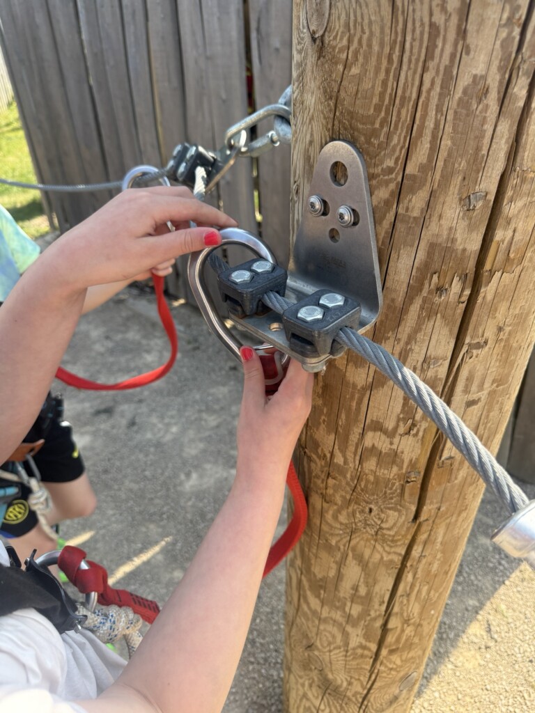Someone fastens a harness and carabiner to a cable on a wooden post at an outdoor adventure course.
