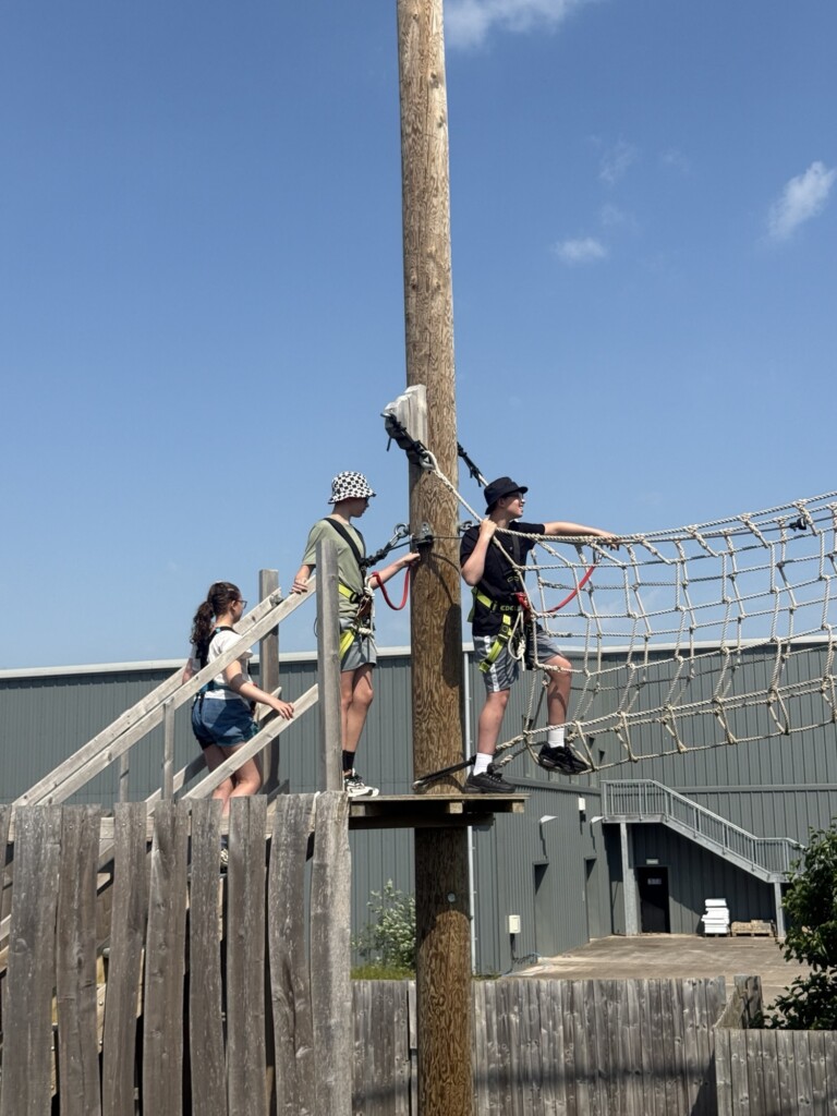 Three people in harnesses stand on a wooden platform by a rope bridge, getting ready to climb outside on a sunny day.