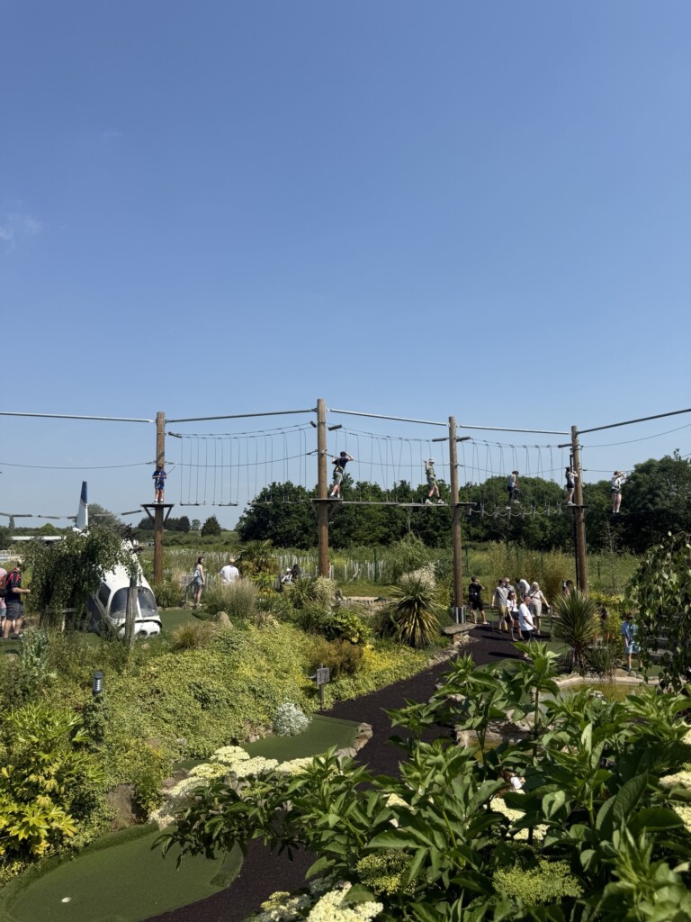 Several people climb a high ropes course outside on a sunny day, with gardens below and some onlookers watching nearby.