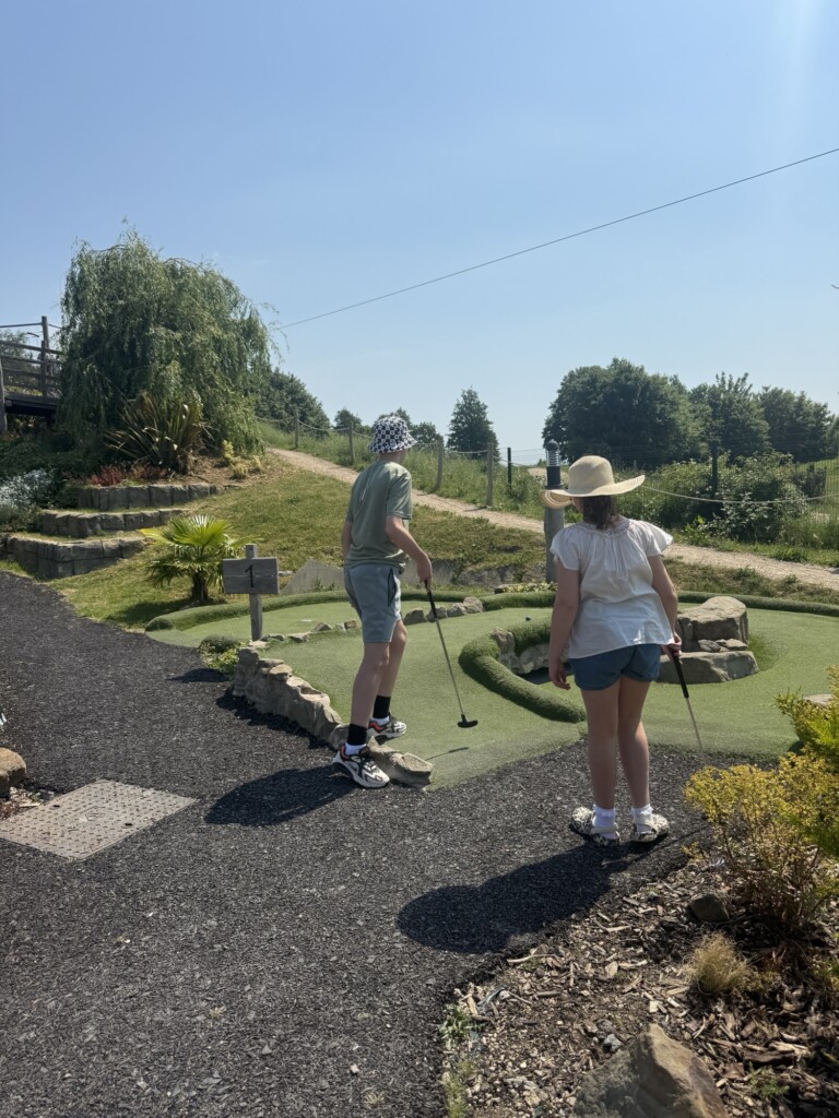 Two kids play crazy golf outside on a sunny day, standing on a green course with plants and landscaping around them.